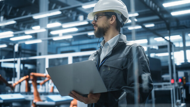 Factory Office: Portrait of Confident Caucasian Male Chief Engineer Wearing Hard Hat Working on Laptop Computer. Technician At Automated Robot Arm Assembly Line Manufacturing Electric Components.