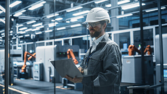 Factory Office: Portrait of Confident Caucasian Male Chief Engineer Wearing Hard Hat Working on Laptop Computer. Technician At Automated Robot Arm Assembly Line Manufacturing Electric Components.
