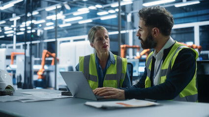 Factory Office: Caucasian Female Quality Control Technician Talks to Hispanic Male Industrial Engineer Working on Laptop Computer. Automated Robot Arm Assembly Line Manufacturing Modern Engines.