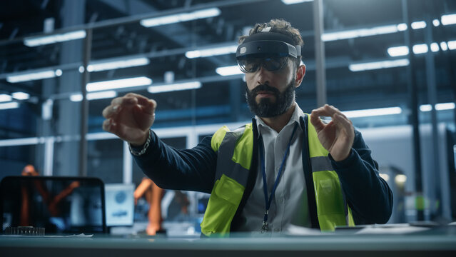 Hispanic Male Industrial Designer Sitting in Factory Office And Using Augmented Reality Headset To Adjust Autonomous Assembly Line With Robotic Arms. Man Gesturing, Using Futuristic Interface.