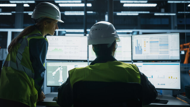 Caucasian Male Engineer Using Multi Monitor Workstation And Talking To Female Production Supervisor At Electronics Factory. Colleagues Monitoring Autonomous Assembly Line With Robotic Arms. Back View