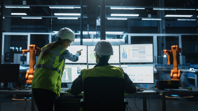 Caucasian Male Engineer Using Multi Monitor Workstation And Talking To Female Production Supervisor At Electronics Factory. Colleagues Monitoring Autonomous Assembly Line With Robotic Arms. Back View - Powered by Adobe