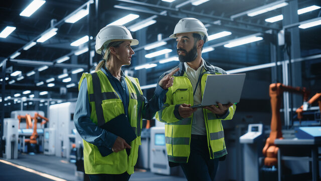 Two Diverse Engineers Walking, Using Tablet Computer at an Industrial Machinery Factory. Caucasian Female and Hispanic Male Specialists Chatting and Monitoring Robots on a Modern Assembly Line.