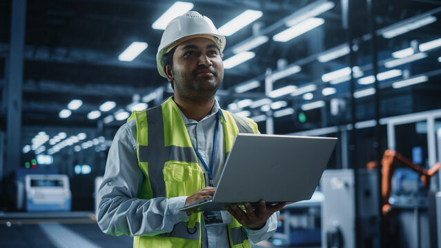 Portrait Of Thoughtful Indian Male Engineer Walking with a Tablet Computer, Monitoring Conditions at an Automated Industrial Machinery Assembly Line with Robot Hands Powered By Artificial Intelligence