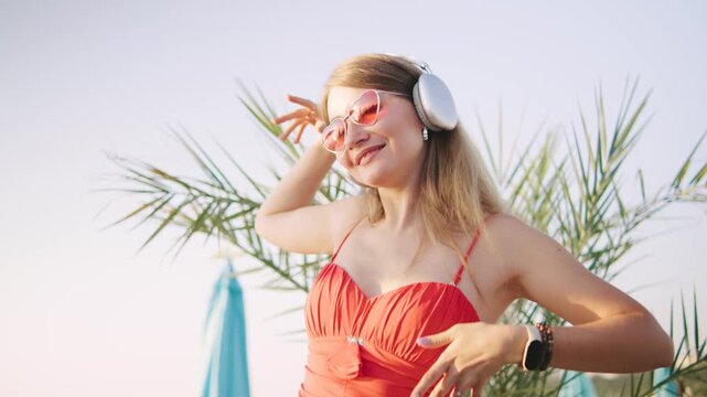 Young blonde woman in headphones enjoys music while dancing at a beach resort on a sunny day surrounded by palm trees and colorful umbrellas