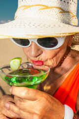 Senior woman in white sunglasses sipping green cocktail with lime garnish. Concept of summer...