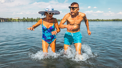Energetic senior couple running through water holding hands on summer beach. Concept of active aging, romantic leisure, wellness, travel joy, summer fun and vitality.