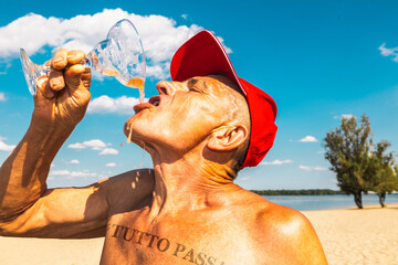 Senior man with drawn Italian phrase Everything Passes on chest, drinking from cocktail glass on beach. Concept of carefree summer moments, vacation relaxation, and playful indulgence.