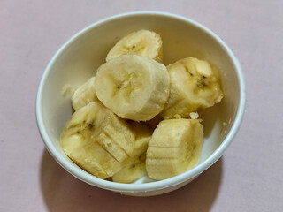 Ripe banana slices served in a white bowl on a plain pink cloth background.