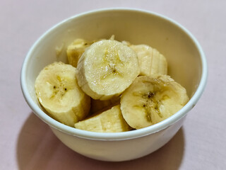 Ripe banana slices served in a white bowl on a plain pink cloth background.