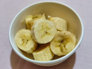 Ripe banana slices served in a white bowl on a plain pink cloth background.