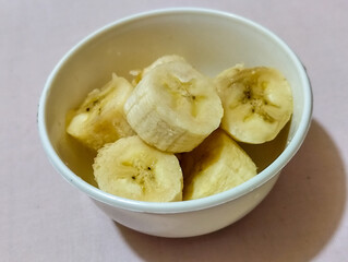 Ripe banana slices served in a white bowl on a plain pink cloth background.