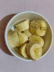Ripe banana slices served in a white bowl on a plain pink cloth background.