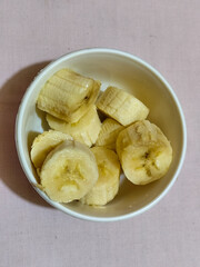 Ripe banana slices served in a white bowl on a plain pink cloth background.