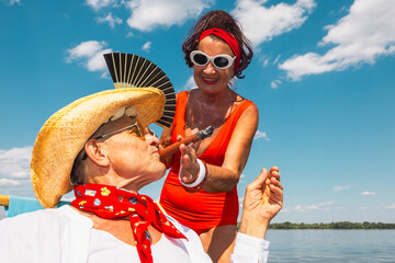 Happy senior couple in summer outfits enjoying cigar by waterfront. Concept of romantic leisure,...