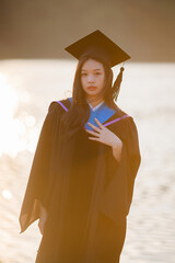 young woman in a graduation cap and gown stands by the tranquil Ang Kaew lake at sunset, looking...