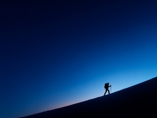 Silhouette of a lone hiker climbing a steep slope at dawn with trekking poles against a deep blue sky