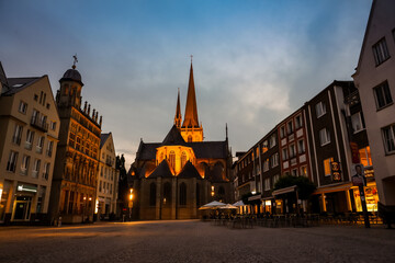 Obraz premium Wesel, Germany – August 6, 2025: Illuminated Willibrordi Cathedral and historic market square at dusk, with surrounding buildings and outdoor cafes.