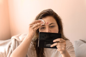 Close-up of a young woman looking at a pimple in the mirror while gently wiping her face with a...