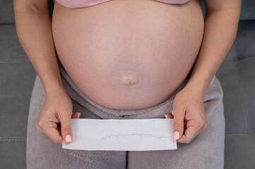A pregnant woman looks at the results of cardiotocography while sitting on the couch. Close up of...