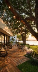 Obraz premium Sunset illuminates a patio restaurant's wood deck beneath a tree strung with lights, overlooking a calm body of water
