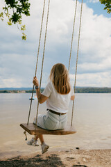 Blonde woman swinging gently on wooden swing above tranquil lake, savoring warmth of relaxing...