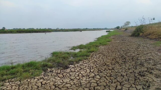 A cracked, dry riverbank meets the calm water under a cloudy sky, illustrating the stark contrast between drought and the remaining source of life.