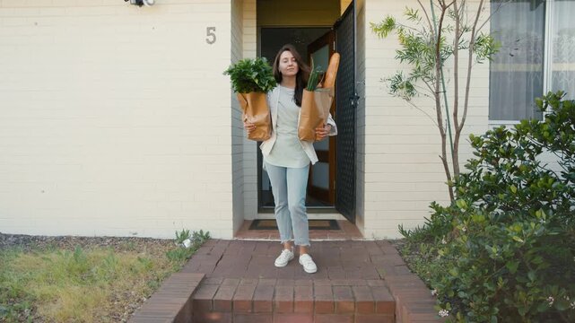 Young adult woman stands at the door of her house holding grocery bags filled with herbs and bread