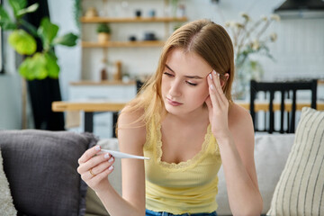 Sad upset teenage girl looking at thermometer sitting at home