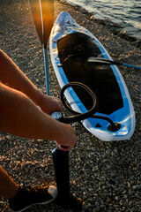 Sportsman inflating sup board with hand pump on pebble beach at sunset, preparing for paddleboarding
