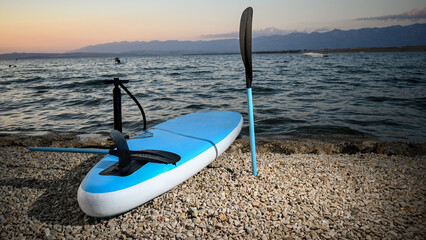 Inflatable paddle board, pump, and paddle lying on a pebble beach at sunset, ready for stand up...