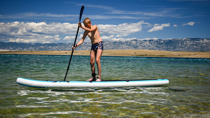 Boy paddling on a stand up paddle board in shallow, clear water with mountain range in the...