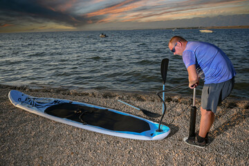 Man inflating his stand up paddle board on a pebble beach at sunset, preparing for watersports