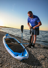 Sportsman inflating sup board with manual pump on pebble beach at sunset, preparing for...