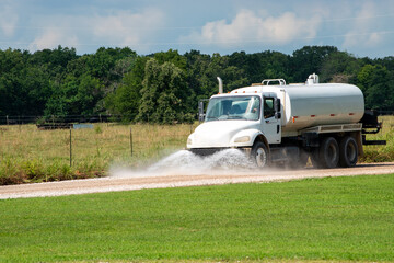 Truck with large tank and powerful pump moves along dirt road as spray nozzels on the front distributes water over the entire road for road maintenance and construction.