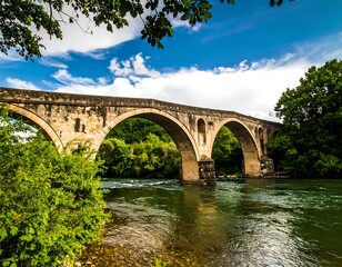 Fototapeta premium Ancient stone bridge over a river, lush greenery