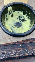 A small black bowl contains a creamy green sauce made from fresh ingredients. It rests on a rustic wooden board, hinting at a meal enjoyed outdoors