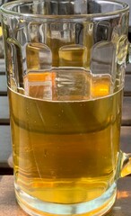 A clear glass filled with a sparkling golden beverage sits on a wooden table. Bright sunlight illuminates the drink, creating a cheerful atmosphere