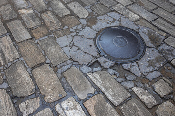 Cast-iron manhole on an old paved road. The road is paved with paving stones. A gray stone is laid...