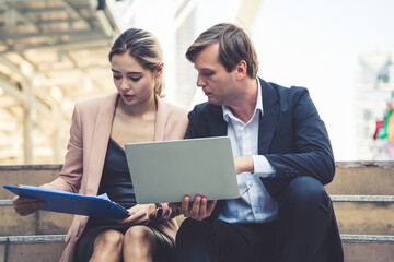 A male and female business colleague work together outside, using a laptop and a document folder. They are focused on their work, collaboration, modern business, and professional partnership.