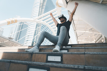 Excited man wearing a VR headset raises his arms in celebration while sitting on urban stairs....
