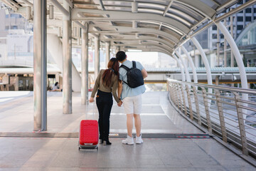 oung Asian couple holding hands and leaning together with a red suitcase in the city. Symbol of love, modern travel lifestyle, digital freedom, and romantic urban tourism journey.