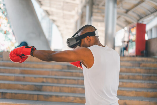 Rear view of a Black male athlete wearing VR headset and red boxing gloves, practicing punches outdoors. Illustrates immersive virtual training, motion tracking, and advanced fitness tech.