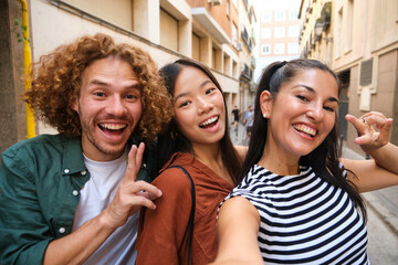 Three happy young multiracial friends are gesturing and taking a selfie in a city street