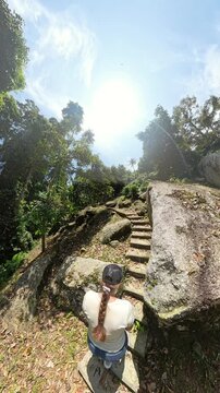 Vertical video. Woman Climbing Over Sunlit Rock Terrain Surrounded by Dense Jungle and Trees with Bright Blue Sky Above, Wide Angle View, Adventure Hiking Concept
