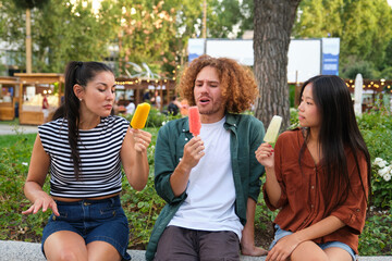 Three friends are sitting on a low wall, tasting different flavored popsicles in a park