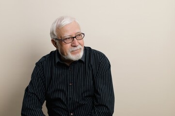 Senior man with glasses smiles softly while seated against a neutral backdrop in a cozy indoor...