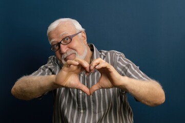 Elderly man makes heart shape with hands expressing love in studio setting on blue background