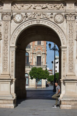 A young, handsome man with a beard, wearing a striped shirt and cargo trousers, rests one leg on a Plateresque and Baroque arch at Seville City Hall in Andalusia. The man is on holiday in the city.