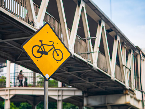 Close-up of a yellow bicycle road sign in front of a pedestrian bridge, highlighting urban infrastructure and cycling safety in the city.